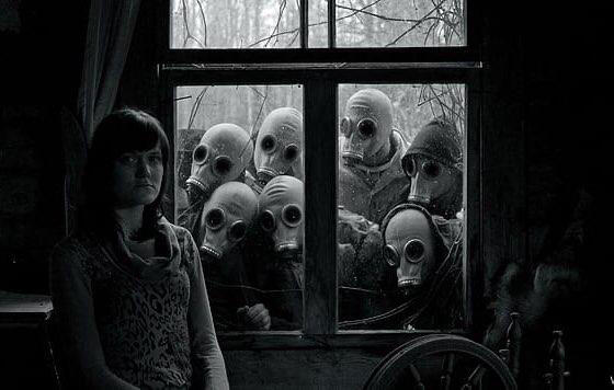 a black and white image of a woman sitting, looking at the camera, a spinning wheel just out of frame, in front of a window where 7 people wearing white gas masks are staring through at her